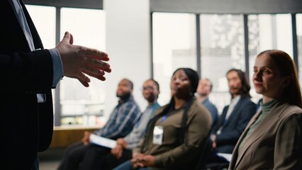 Business summit in conference hall with professional audience seated on chairs. Speaker delivers presentation during panel discussion, encouraging engagement and discussion among attendees.