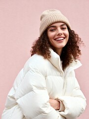 Happy smiling woman in winter fashion. Young female model wearing a beige beanie and a white puffer jacket against a pink background