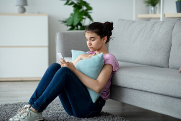 Stressed Indian teen girl sitting on floor with cellphone and reading bad message at home. Upset...