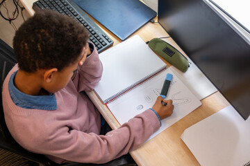 Young student concentrates while writing in a notebook, doing homework at a desk with a computer...
