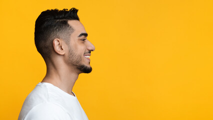 Profile portrait of cheerful bearded arab guy smiling and looking at copy space over yellow studio background, panorama. Handsome middle-eastern young man watching nice advertisement © Prostock-studio