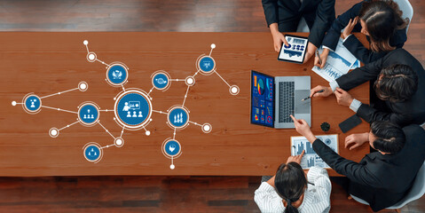 A dynamic overhead view of a business meeting featuring diverse professionals engaged in discussion and analyzing digital data on a laptop and tablets in a modern workspace. Amity
