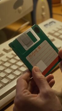 Vertical close-up of anonymous female hands holding vintage 3-inch green floppy disk with work project files, report or document, sitting at desk with retro computer