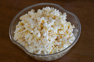 A bowl of freshly popped microwave popcorn in a clear bowl placed on a brown wooden table top.