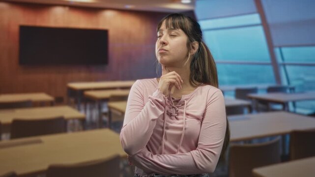 Young hispanic woman with hand on chin among empty classroom desks in a modern lecture building, looking up in pensive pose; thoughtful study.