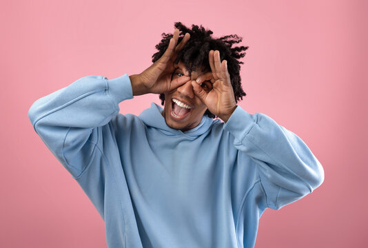 Silly black teen guy doing airplane glasses with his fingers, grimacing, making funny face on pink studio background. Playful African American adolescent joking, being carefree and comical