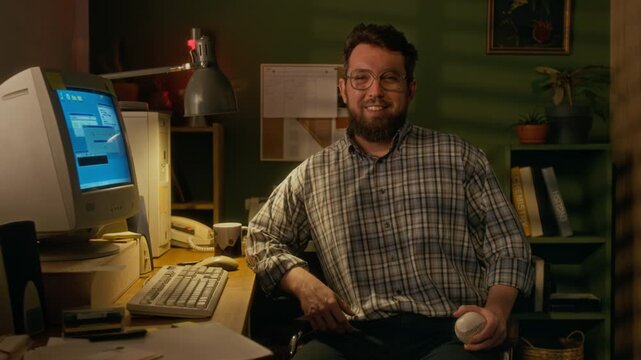 Medium portrait of cheerful young Caucasian male programmer sitting at desk next to vintage desktop computer with CRT monitor and tower case while working from home, looking at camera, smiling
