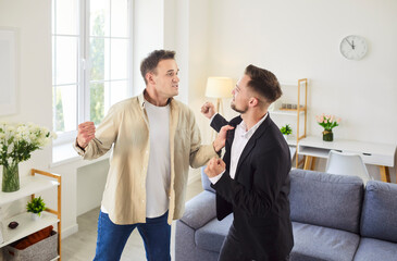 Two furious men face each other in a tense argument in a living room. Angry opponents have raised fists, shouting and threatening physical fight. The atmosphere is charged with tension.