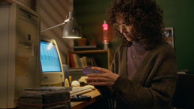 Medium shot of young Caucasian woman in glasses, with curly hair inspecting CD disks, checking surface for marks, then putting in box, while working on report on old-fashioned computer at home