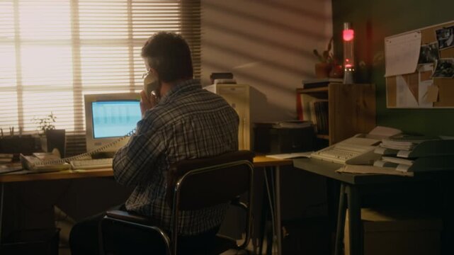 Medium rear backlit shot of Caucasian businessman working on old computer, with printer and fax machine, answering call using corded telephone at cozy home office with window blinds