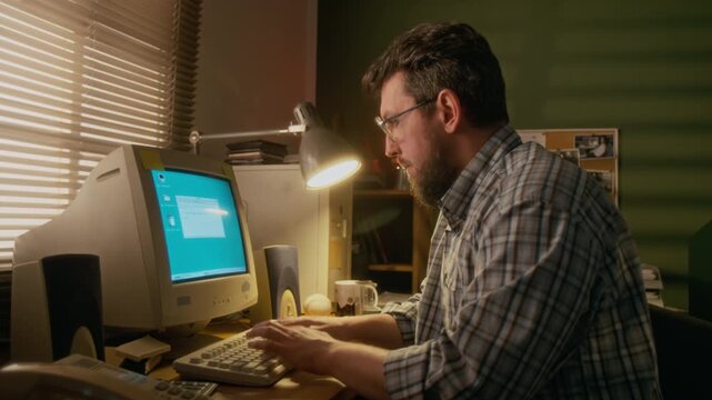 Medium shot of Caucasian male manager sitting at desk at home study, typing on keyboard of vintage computer with flickering CRT screen, getting email from client and nodding with satisfaction