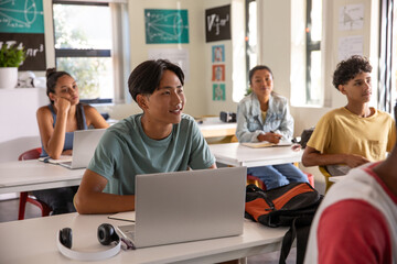 Teenage male student sitting at front desk in class using silver laptop and white headphones