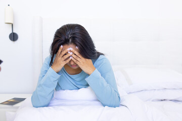 Asian woman leaning forward on bed wearing pale blue top, covering face, bedside table holding book