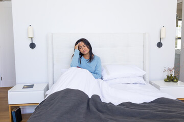 Asian woman sitting on bed, holding forehead, wearing light blue top, closed book visible