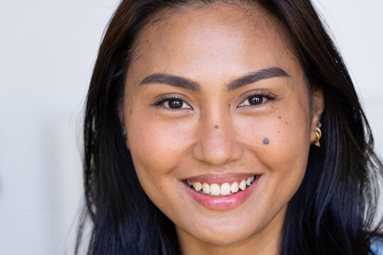 Mid adult Asian woman smiling, showing beauty marks, wearing gold hoop earring, dark top in studio