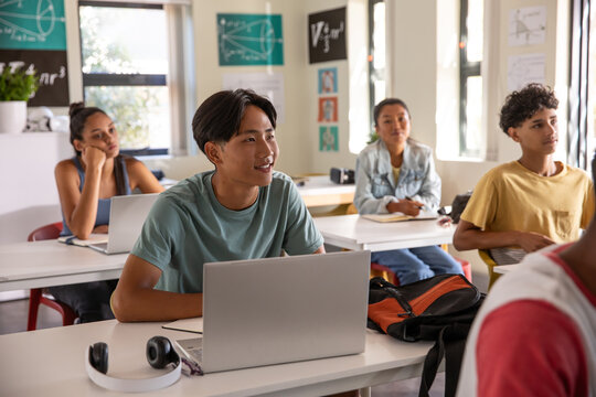 Teenage male student sitting at front desk in class using silver laptop and white headphones - Powered by Adobe