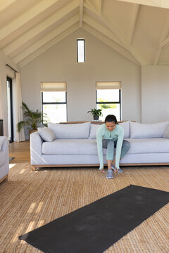Mid-adult Asian woman in green top, leggings tying shoes on sofa in living-room with yoga mat