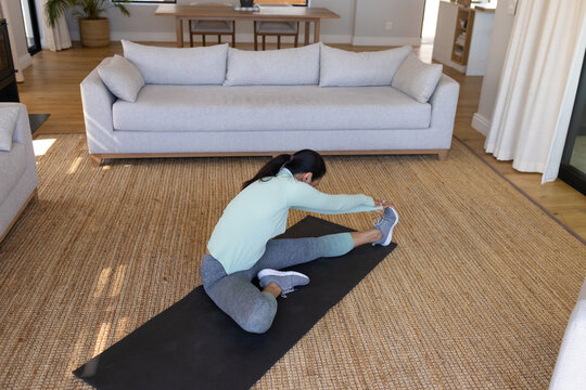 Asian woman stretching on black yoga mat on jute rug in living room, in athletic clothes