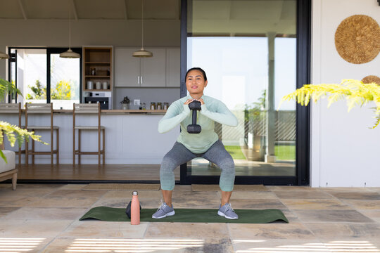 Asian woman squatting on green mat on patio holding dumbbell with pink bottle in leggings