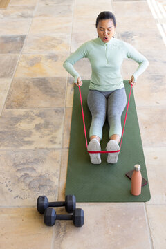 Asian mid adult woman pulling resistance band, seated on green mat on tiled floor with dumbbells