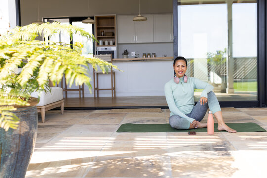Mid adult Asian woman sitting on yoga mat at tiled patio, pink bottle, headphones, copy space