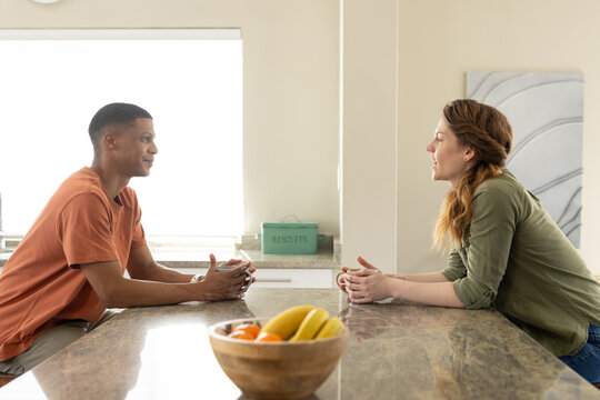 Diverse couple leaning on kitchen island, sipping from mugs near fruit bowl and BISCUITS tin
