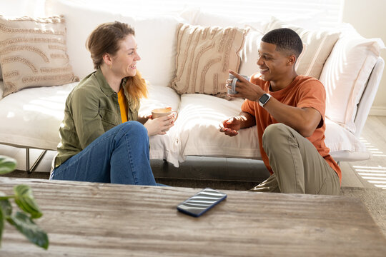 Diverse friends sitting on floor in sunlit living room, holding ceramic mugs, smartphone visible