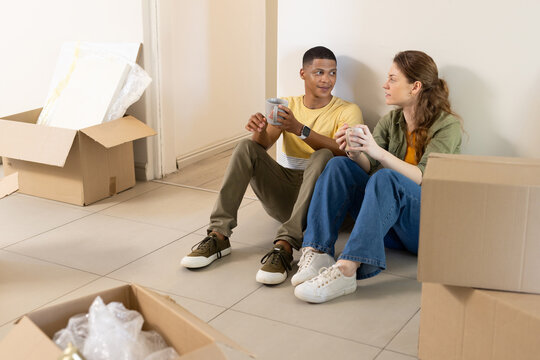 Couple sitting on tiled floor in empty room, holding ceramic mugs near bubble wrap and boxes