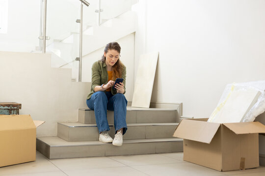 Woman sitting on tiled stair landing surrounded by open moving boxes, checking smartphone