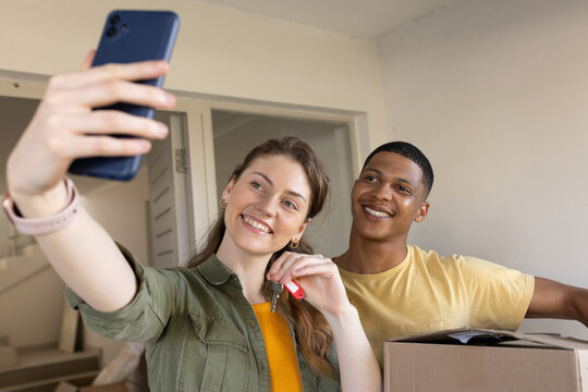 Diverse couple taking phone selfie holding house key with red cover in entryway amid moving boxes