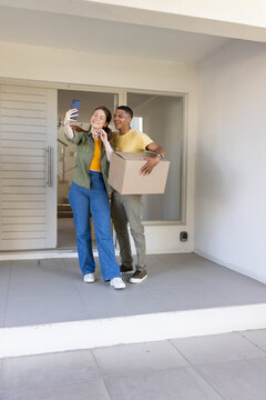 Couple taking smartphone selfie on tiled front porch with man holding box and woman holding keys