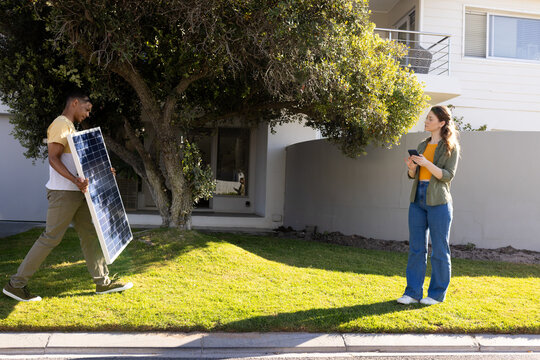 Diverse pair walking on lawn, carrying solar panel and holding tablet, copy space