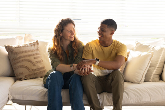 Couple sitting close on light-colored sofa in living room, holding hands, wearing smartwatches