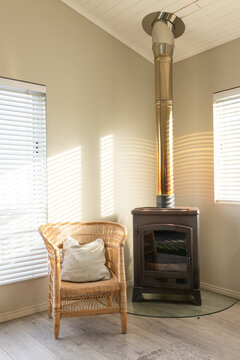 Metal wood stove is sitting on glass hearth pad in corner, armchair and blinds casting stripes