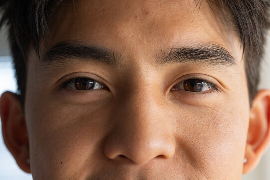 Asian man looking into camera in room near window, showing brown eyes and skin texture