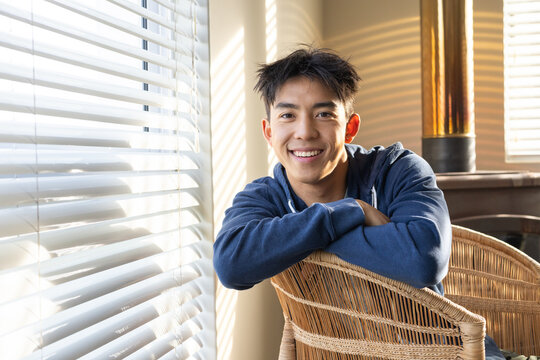 Adult Asian man sitting on wicker chair by window with white horizontal blinds, wearing blue hoodie