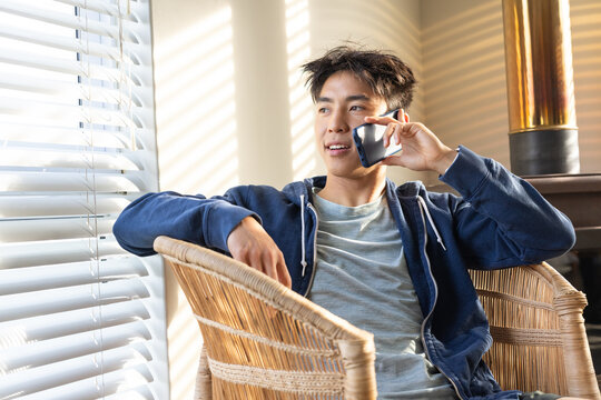 Adult Asian man sitting in wicker armchair by large window with blinds, holding smartphone