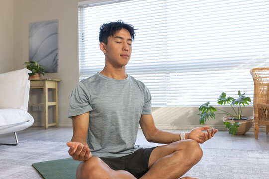 Adult Asian man sitting cross-legged meditating on green yoga mat in living room wearing gray tee