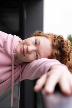 Female leaning, resting head on arm on metal railing on balcony in pink hoodie with drawstrings