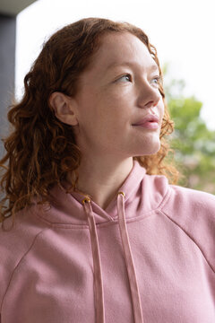 Woman gazing upward to right, wearing pink hoodie with metal eyelets by porch column and trees