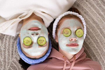 Diverse female friends lying head-to-head on grey rug, wearing clay masks with cucumber slices