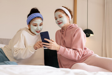 Diverse female friends sitting on bed in bedroom, wearing hoodies, green masks, holding phone