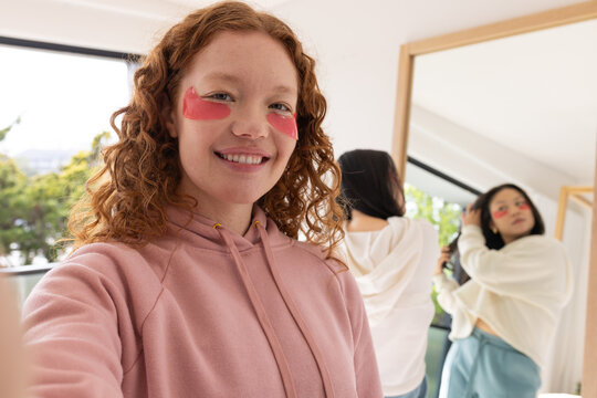 Diverse female friends taking selfie wearing pink hoodie red under-eye patches styling hair at home