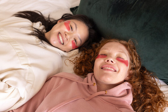 Diverse female friends lying on bed in bedroom wearing red under-eye patches and hoodies