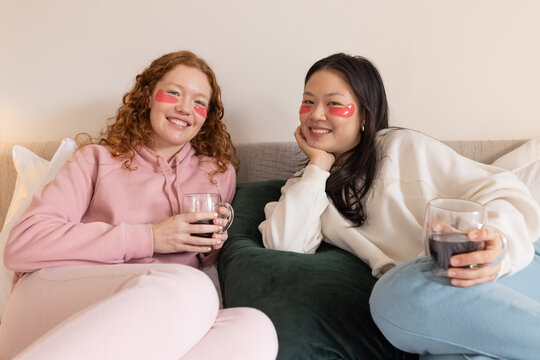 Diverse female friends lounging on beige sofa at home wearing sweats pink eye patches holding glass