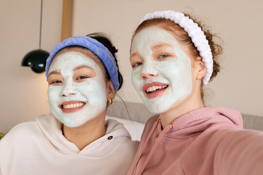 Diverse female friends posing for selfie on couch wearing clay masks, hoodies, terry headbands