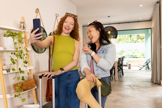 Diverse female friends posing for selfie at rental entry, holding smartphone and rolling suitcases