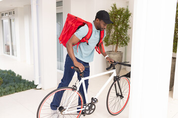 African American mid adult man guiding white bicycle across tiled home porch wearing red backpack