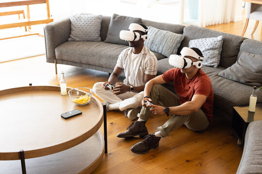 Diverse male friends gaming wearing white VR headsets and holding controllers on hardwood floor