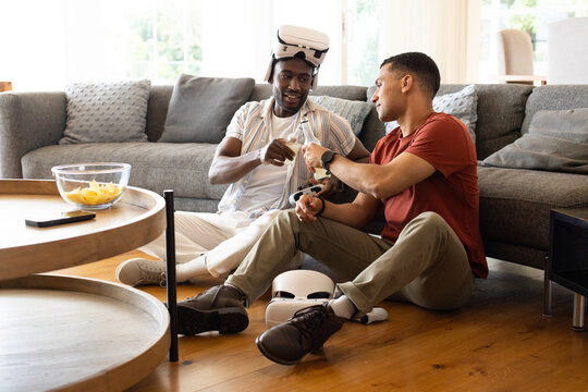 Diverse male friends sitting on hardwood floor in living room clinking bottles near VR headsets